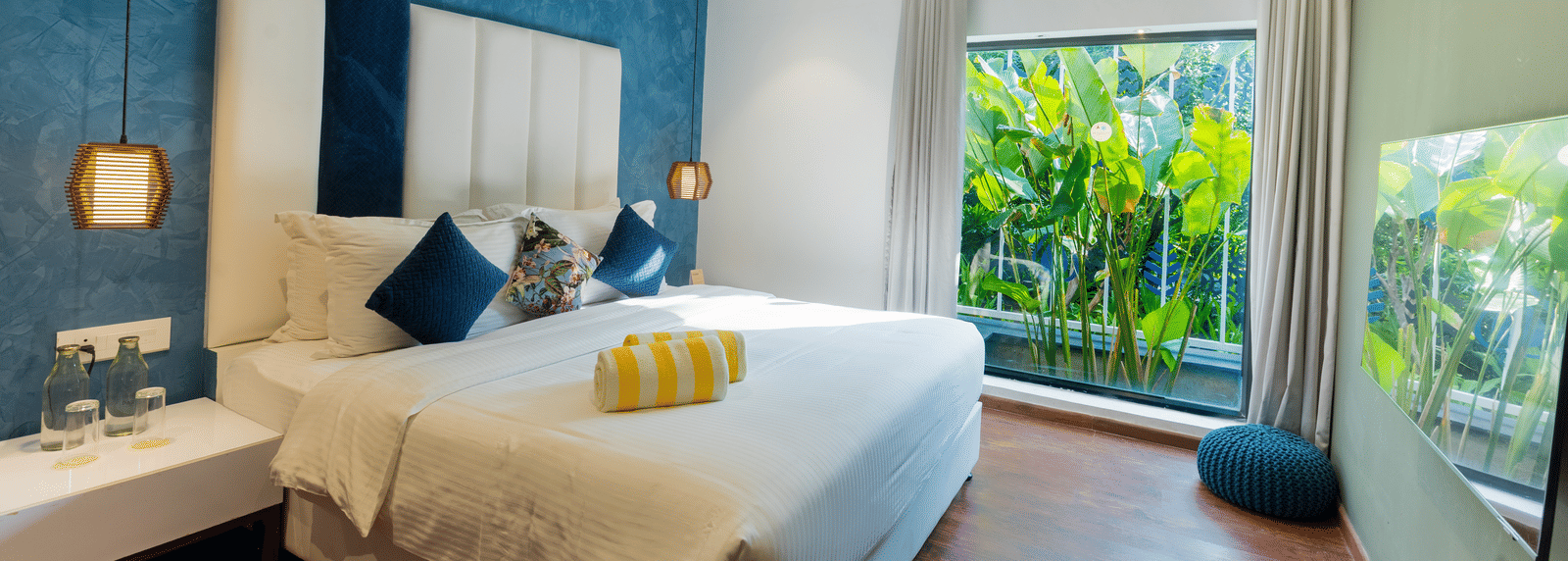 Bedroom of Two Bedroom Villa at Darza Resorts features a large bed, blue accent wall, and a full-length window overlooking lush tropical foliage.  - MICE Tourism in Coimbatore