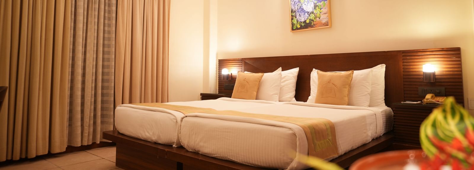 A warmly lit Superior Room at Vedic Village Spa Resort featuring a double bed with a wooden headrest, drapes over a large window, and a painting above the bed.