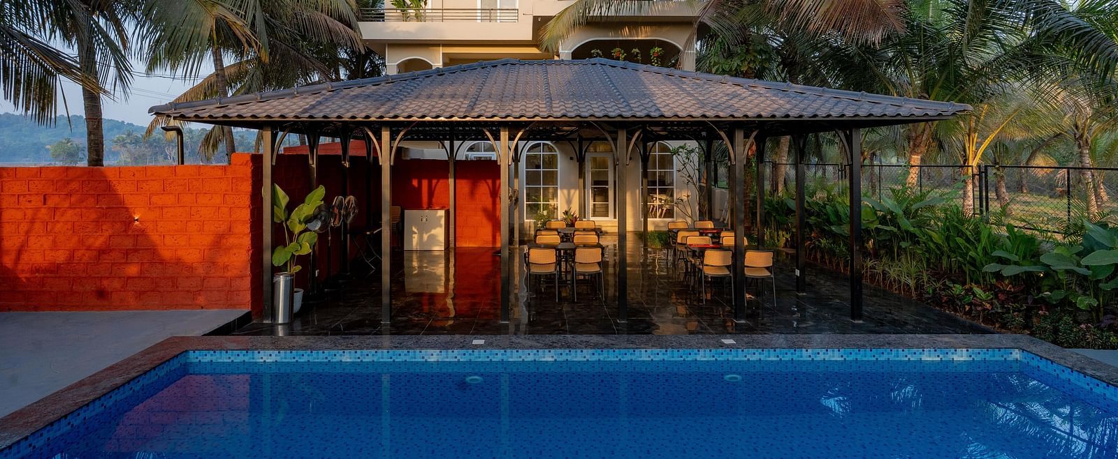 A view across a swimming pool towards a covered outdoor seating area with a thatched roof. Palm trees sway gently in the background at Kssemya.