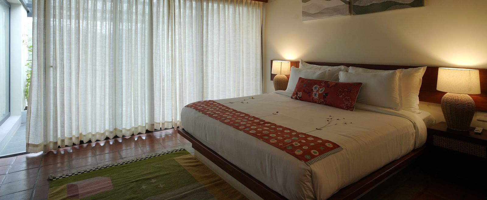 A deluxe room at Vaasta Vagamon featuring a large bed with decorative throw pillows and a patterned runner, and long drapes.