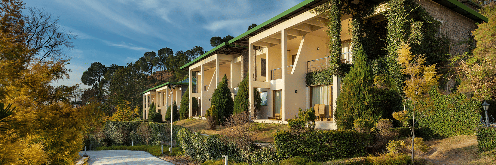Facade of Suryavilas Luxury Resort and Spa in Solan with a paved driveway and green climbing plants on its exterior walls, under a blue sky with clouds