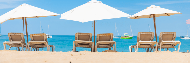 A row of empty loungers under white umbrellas lines a serene beachfront at The Soco Hotels, facing the clear blue sky and calm ocean.