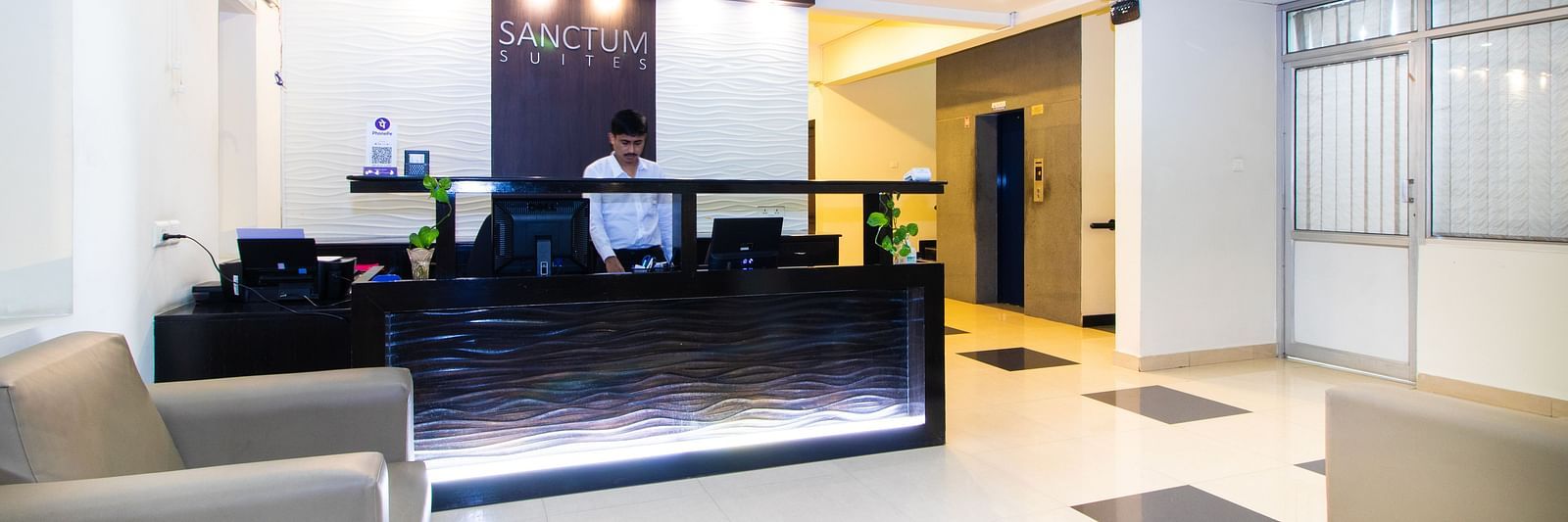 A lobby area with seating, a sleek reception desk, and modern lighting fixtures at Sanctum Suites Indiranagar, Bangalore.