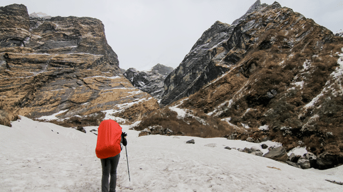 A hiker walking through a snow-covered mountain trail with rocky slopes.