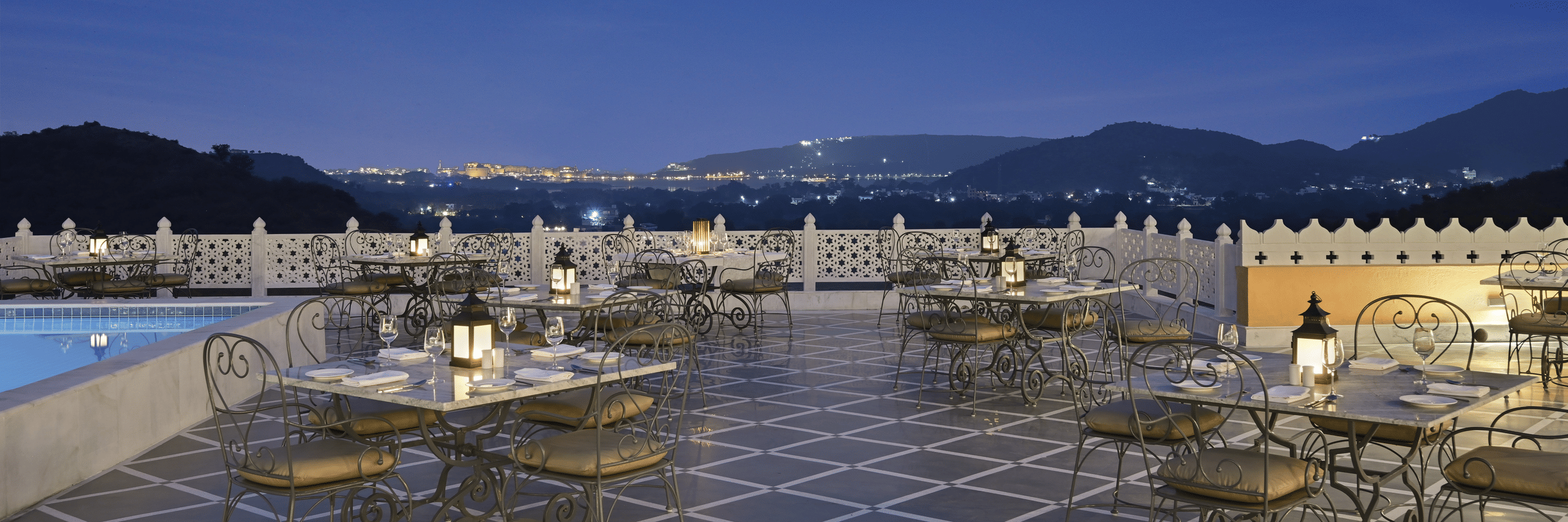 Terrace next to the swimming pool with seating arrangement for fine dining in Udaipur at Fateh Vilas and views of the mountains in the twilight sky
