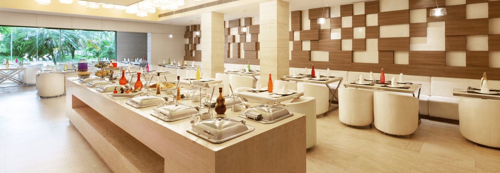 A diagonal view of a restaurant with an island in the middle featuring buffet items, surrounded by tables and chairs on the sides | Cenneys Gateway