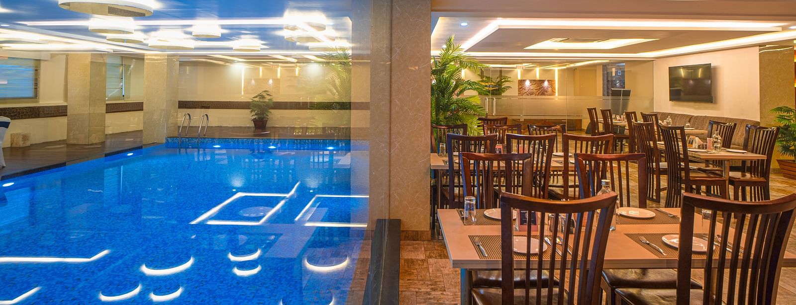 Poolside dining area with chairs, tables, and ceiling lights beside the indoor pool at Mansha Regency, Port Blair.
