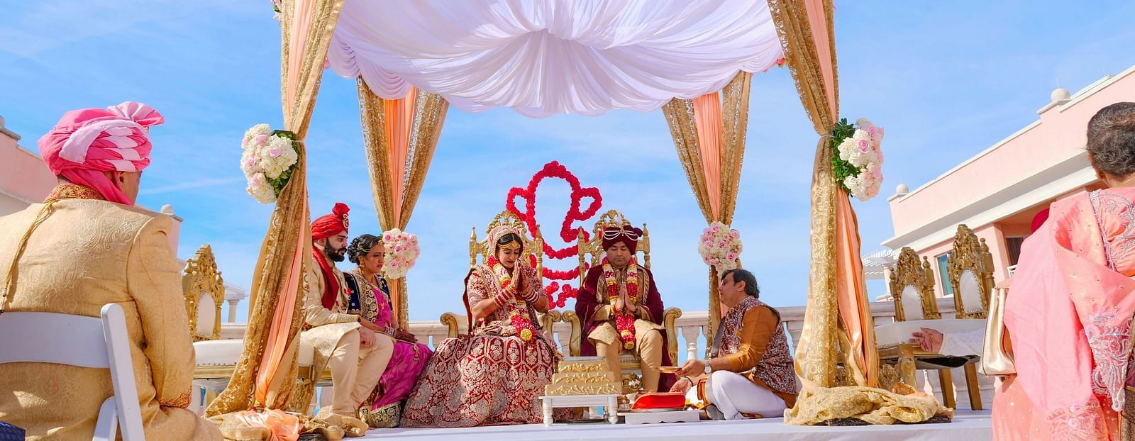This image showcases a grand Indian wedding ceremony taking place outdoors under a beautiful, floral-adorned setup. 