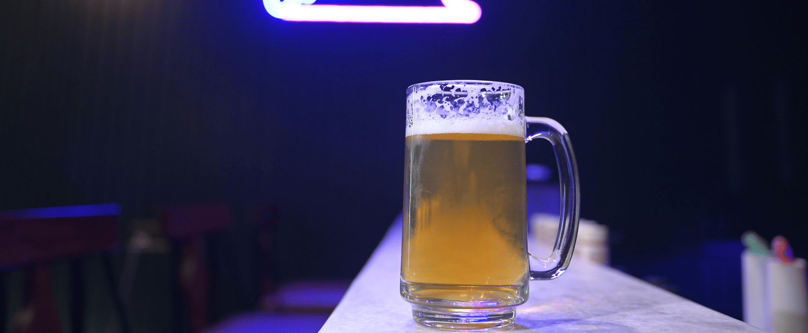 A chilled beer mug placed on the counter with a glowing neon cocktail sign in the background at Hotel Sonar Bangla Mayapur.