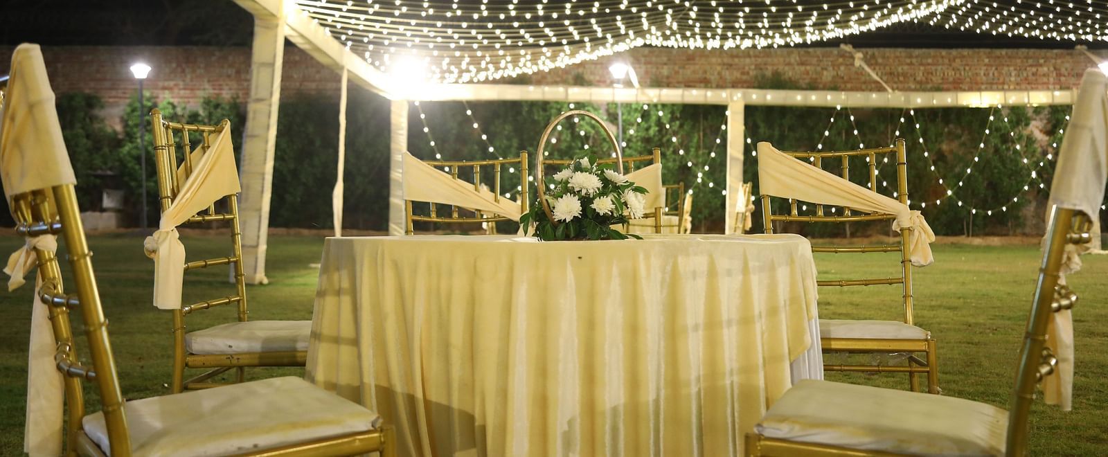 An outdoor seating area at Tisya Farms, Gurgaon with string lights overhead, a striped tablecloth, and white drapes.