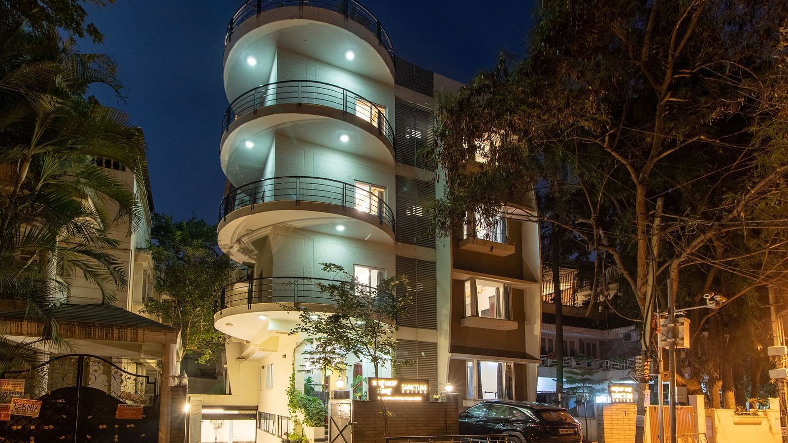 An exterior view of Sanctum Suites Indiranagar, Bangalore showcasing the cylindrical building illuminated at night along with greenery around.
