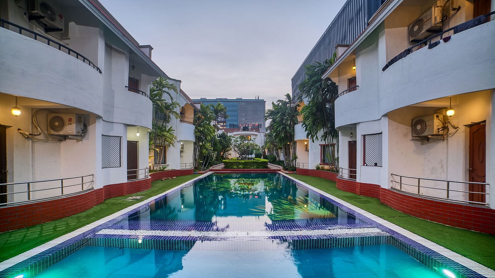A symmetrical view of Ilara Hotels & Spa, OMR, Chennai illuminated at dusk, lining a long, reflective swimming pool or water feature.