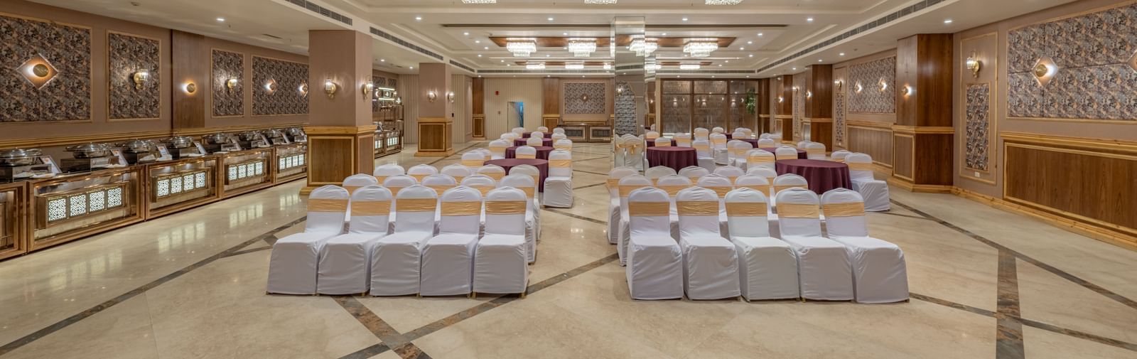Large banquet hall at Hotel Highway King, Bagru, with rows of chairs, decorative ceiling lights, and mirrored wall panels.