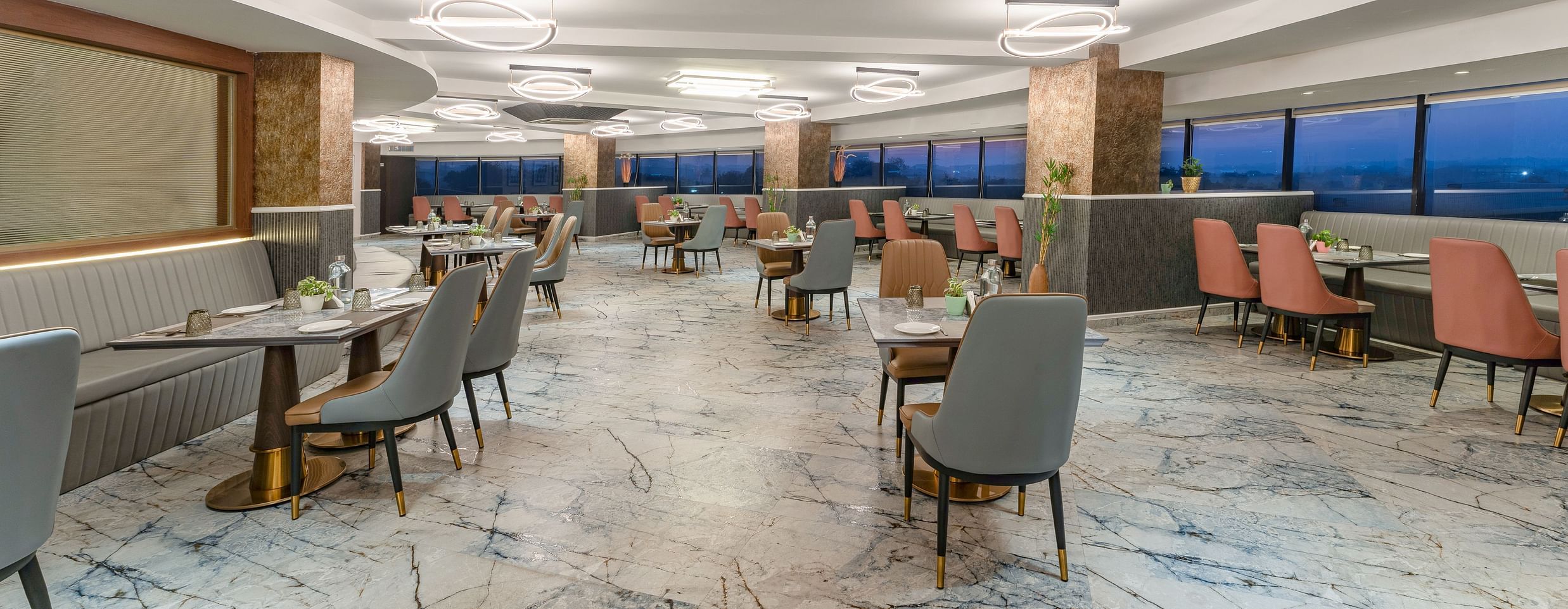 Spacious restaurant interior featuring marble flooring, grey and terracotta chairs, panoramic windows overlooking city skyline at Clarks Inn Suites, Hyderabad