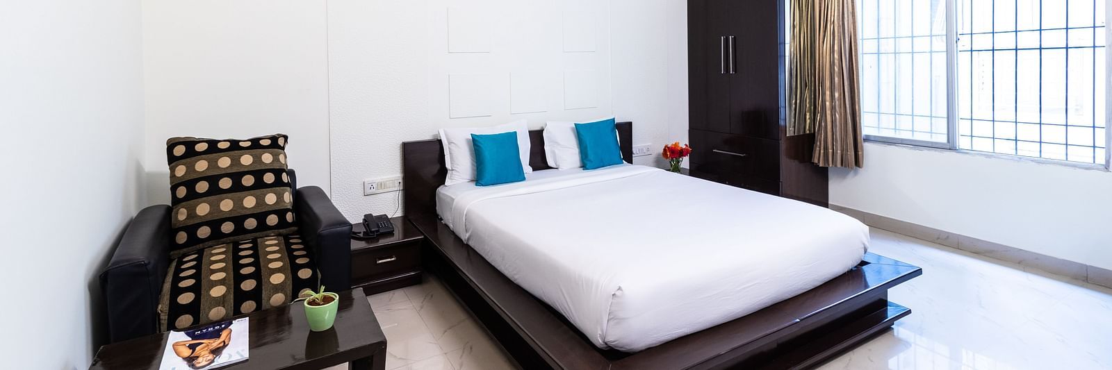 A room with a bed, an armchair, a wardrobe, and a large window at Sanctum Suites Domlur, Bangalore.