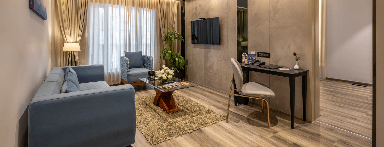 A side view of a living area in the Citrine Hotel, showing a sofa, a chair, a work desk and a wall-mounted TV.