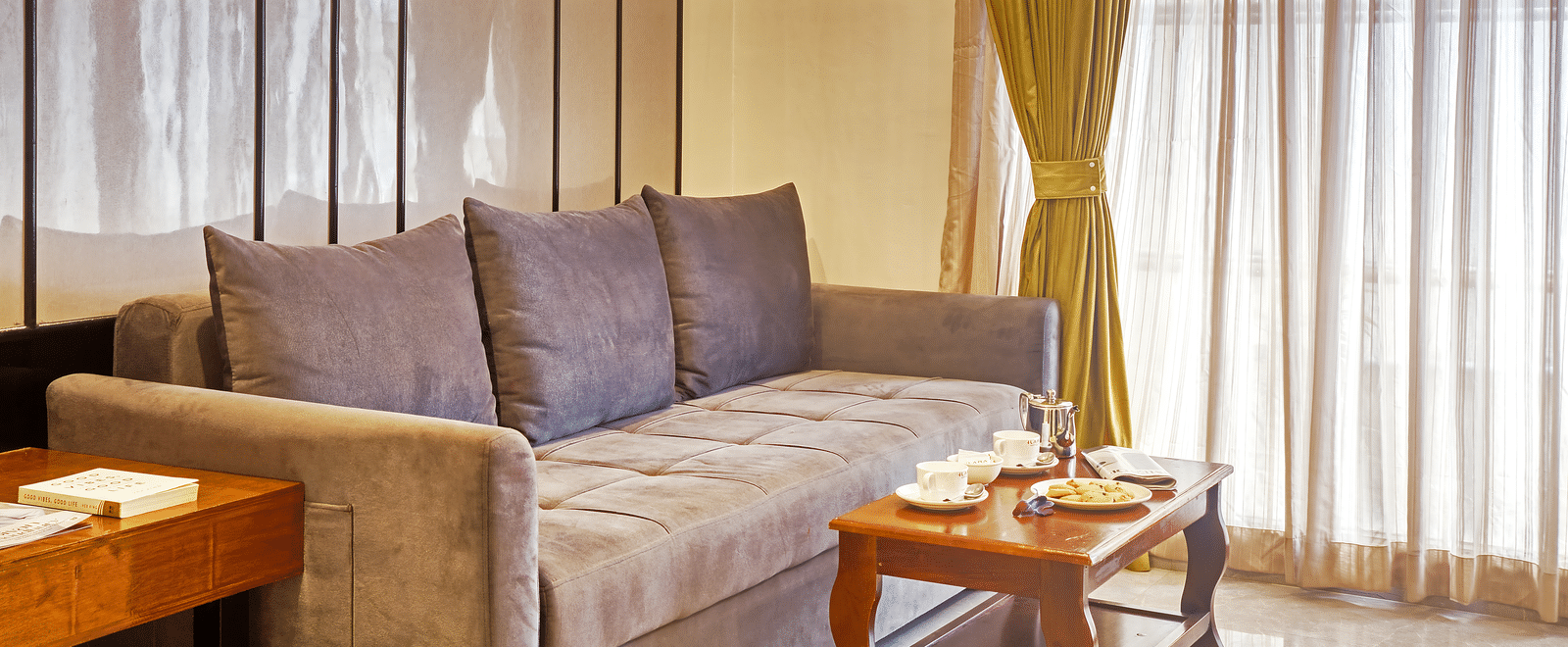 Living room with grey sofa, sheer curtains, and coffee table in a well-lit space at Ilara Hotels & Spa, OMR, Chennai