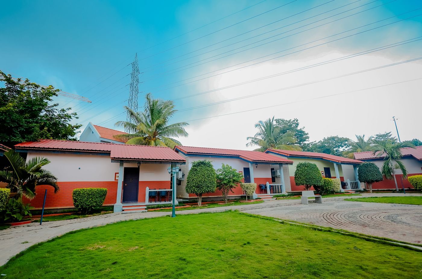Exterior view of cottages with red roofs and green lawns at Nirantara Resort & Spa, Bengaluru, under a partly cloudy sky.