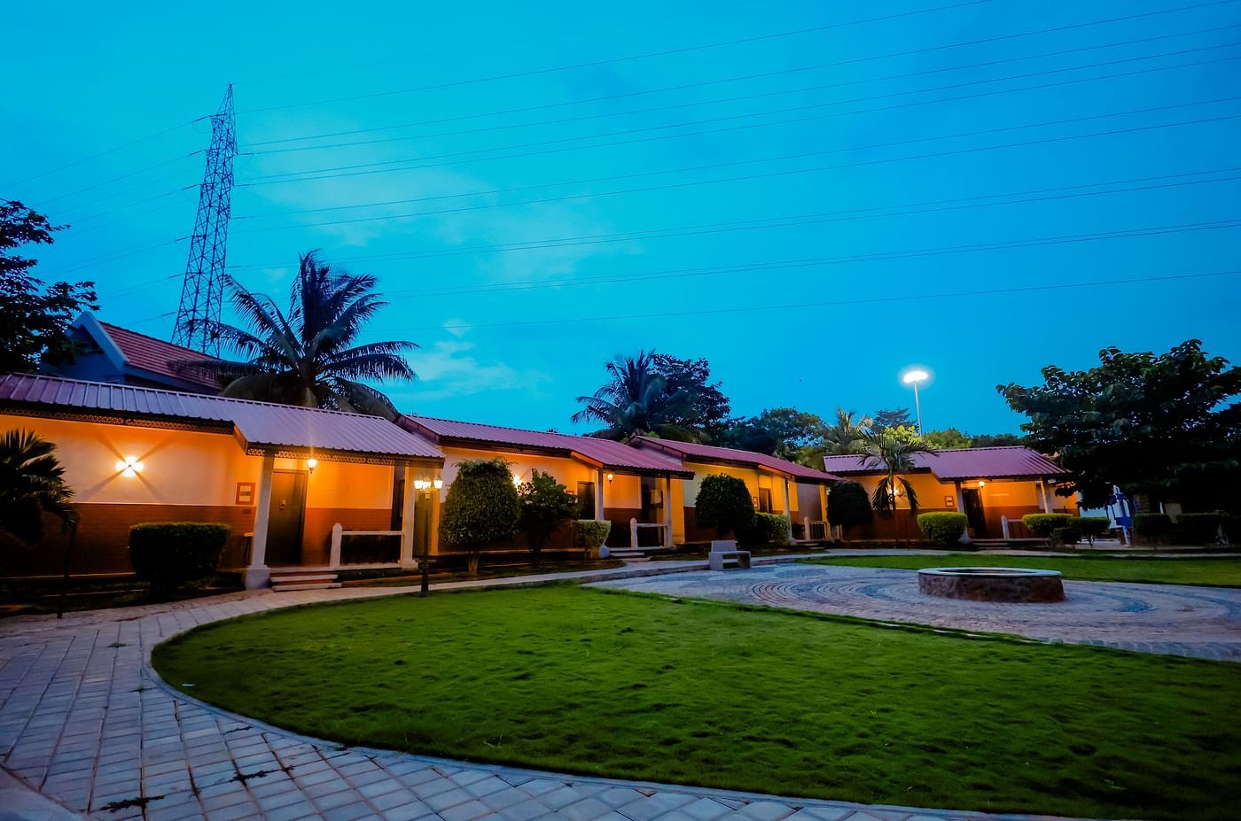 Exterior view of cottages with red roofs and green lawns at Nirantara Resort & Spa, Bengaluru, at dusk with lights illuminating the pathways.