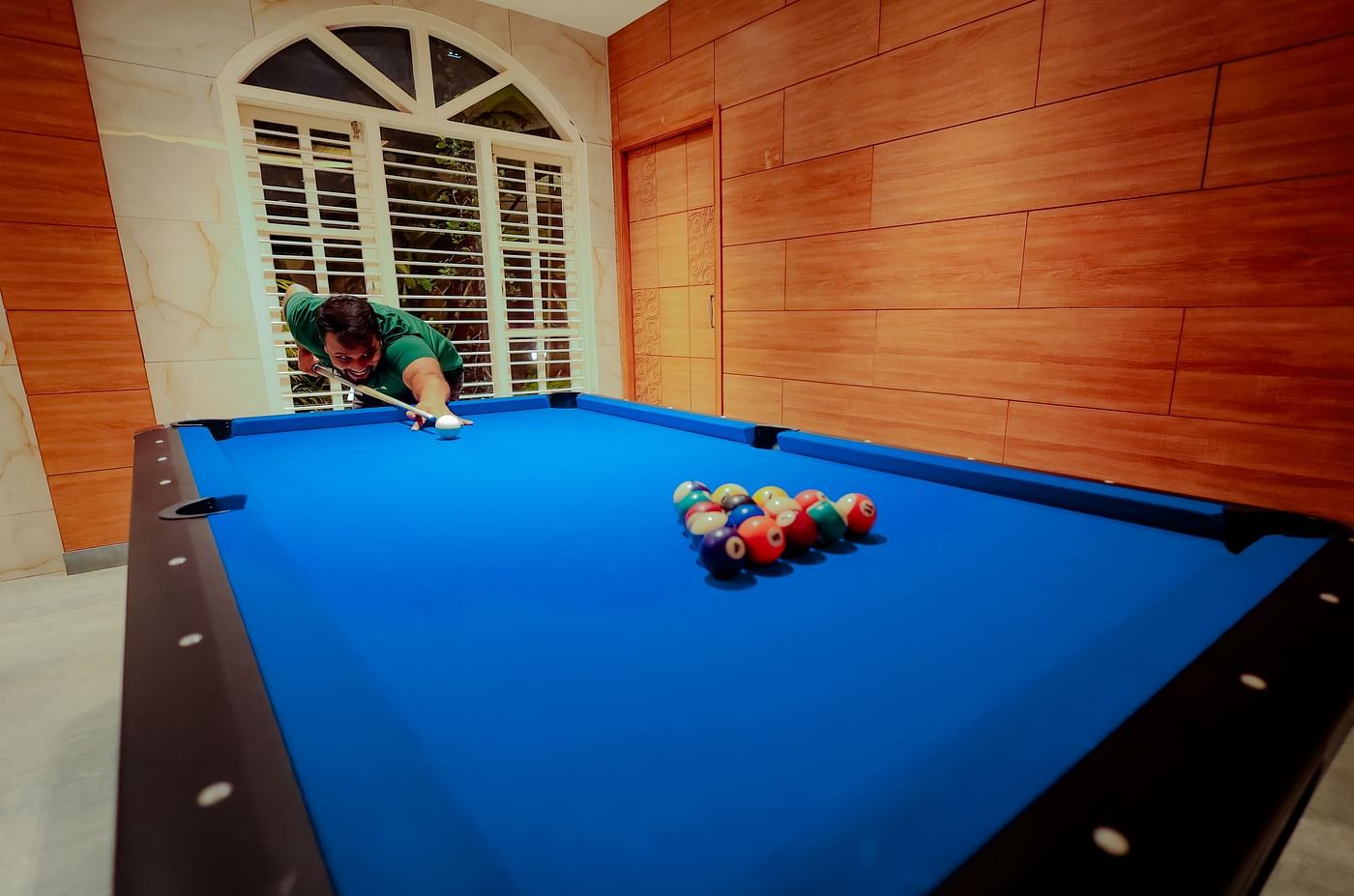A person playing pool on a blue pool table in a room with a brick wall and arched window at Nirantara Resort & Spa.