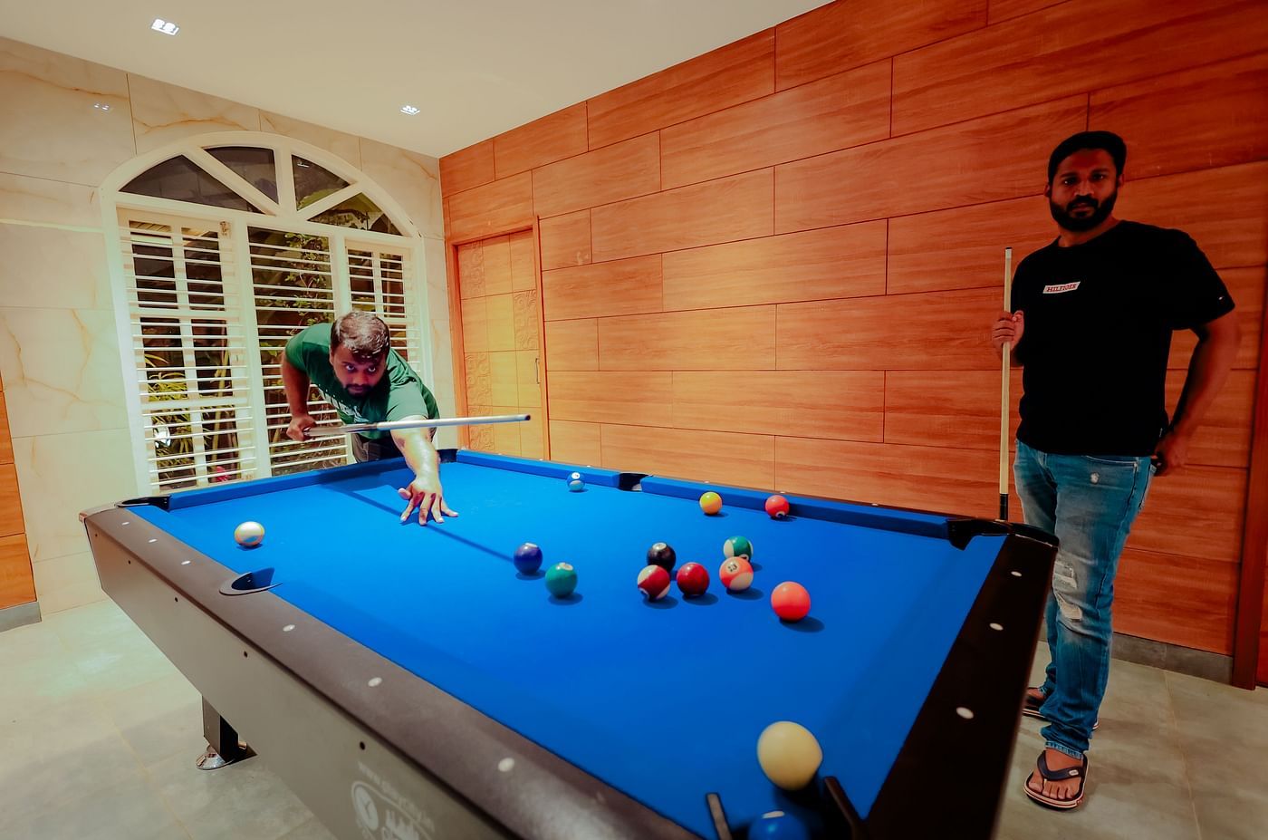 A person plays pool on a blue pool table in a room with brick walls and large windows - Nirantara Resort & Spa.