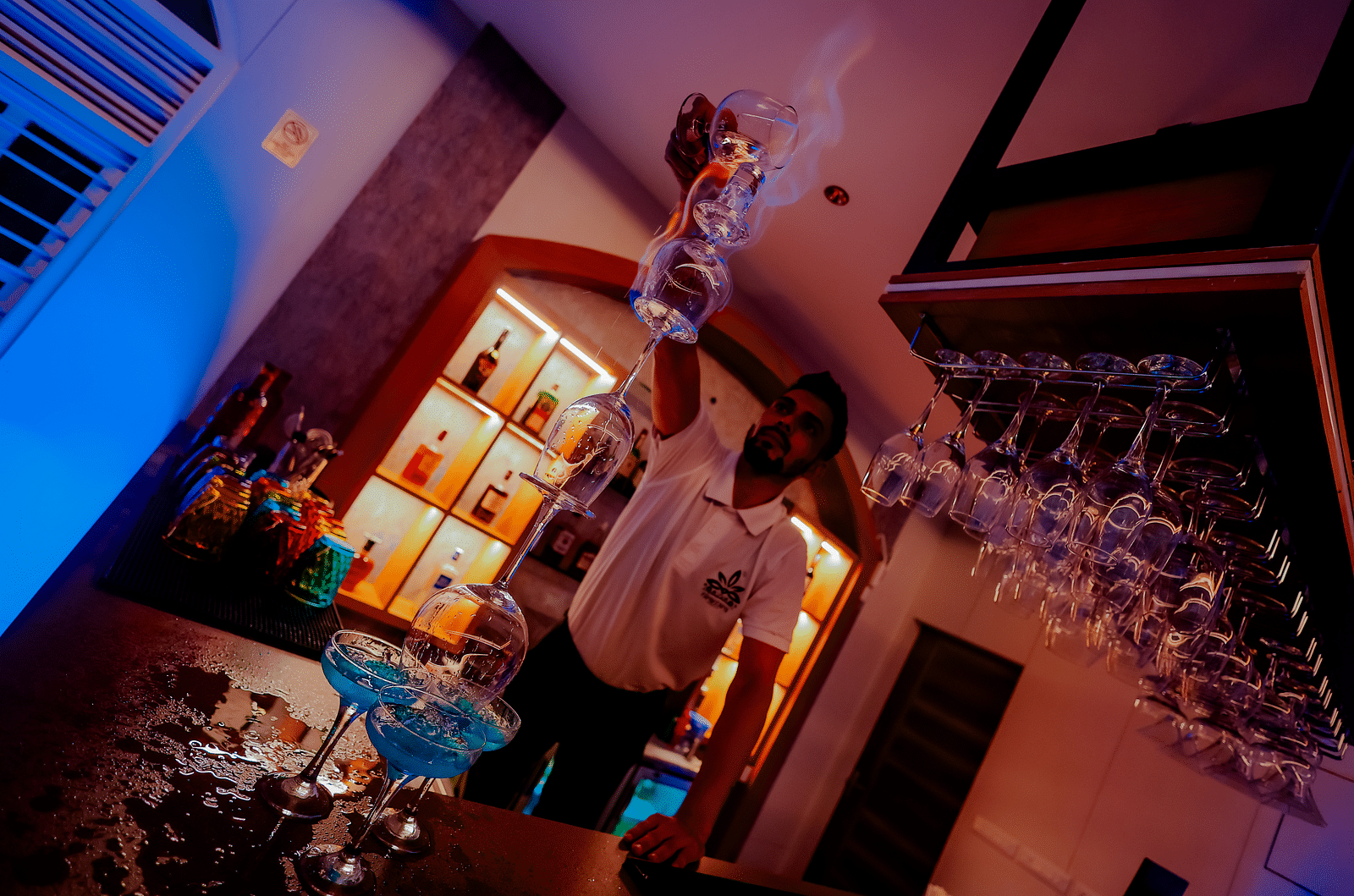 Low angle shot of a bartender pouring a drink into a glass, with rows of glasses and bottles visible in the background - Nirantara Resort & Spa.