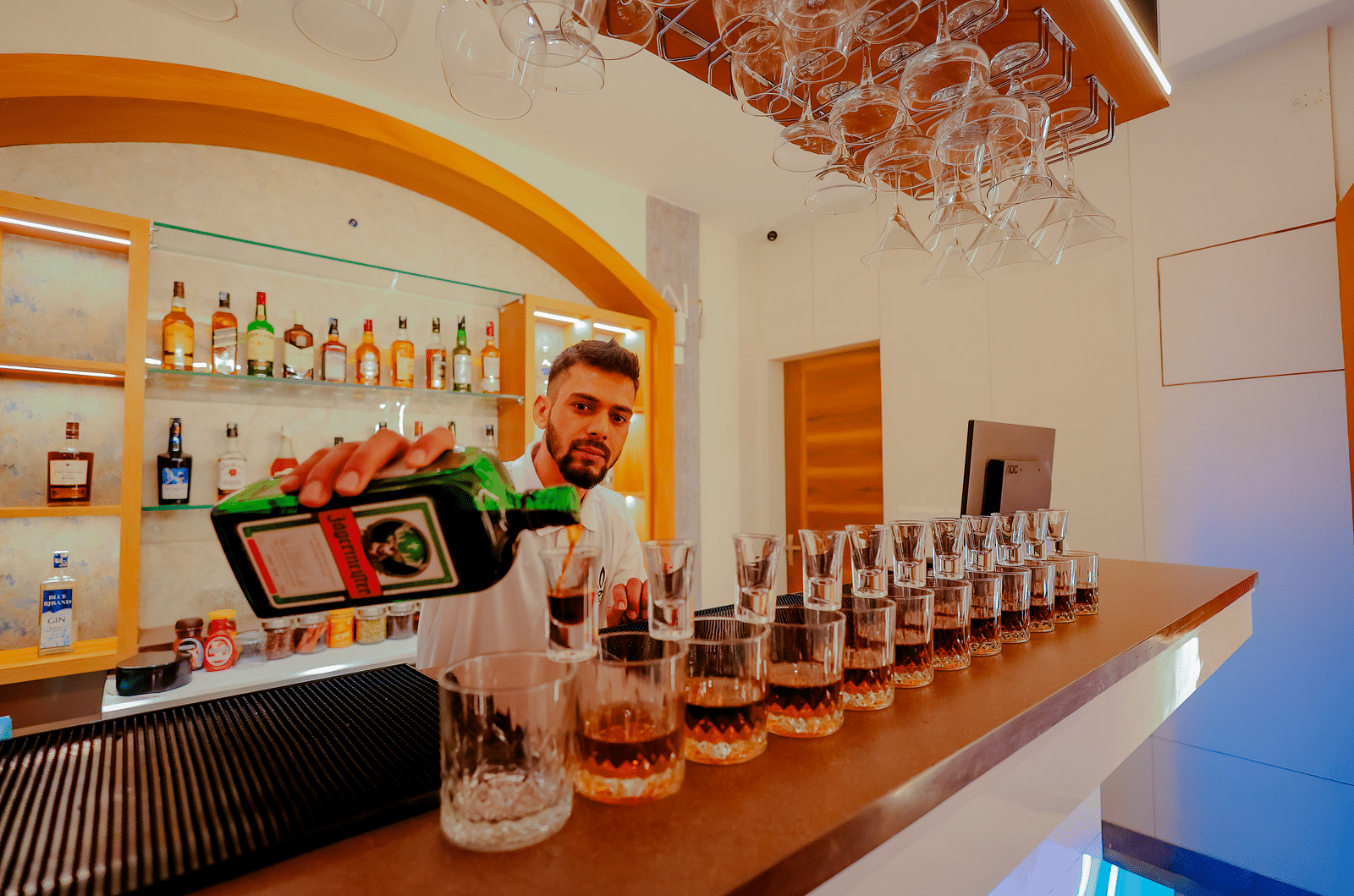 A bartender prepares drinks at a well-stocked bar at Nirantara Resort & Spa, showcasing a variety of liquors and glassware.