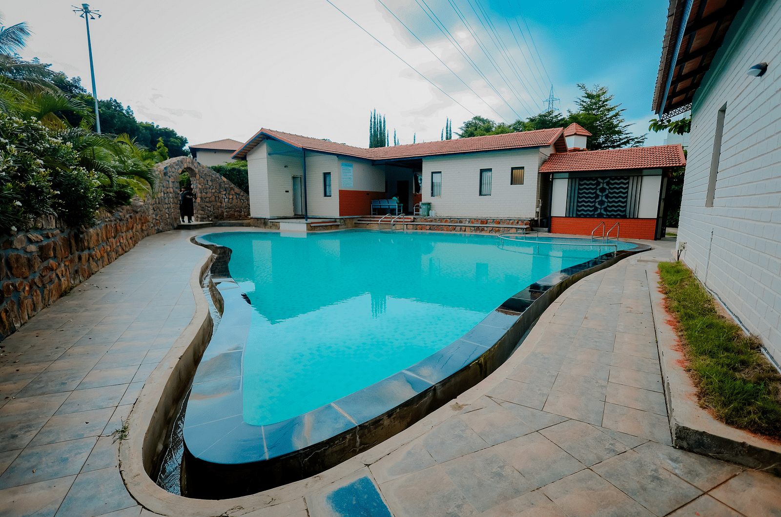 A uniquely shaped swimming pool at Nirantara Resort & Spa, Bengaluru, surrounded by plants and buildings.