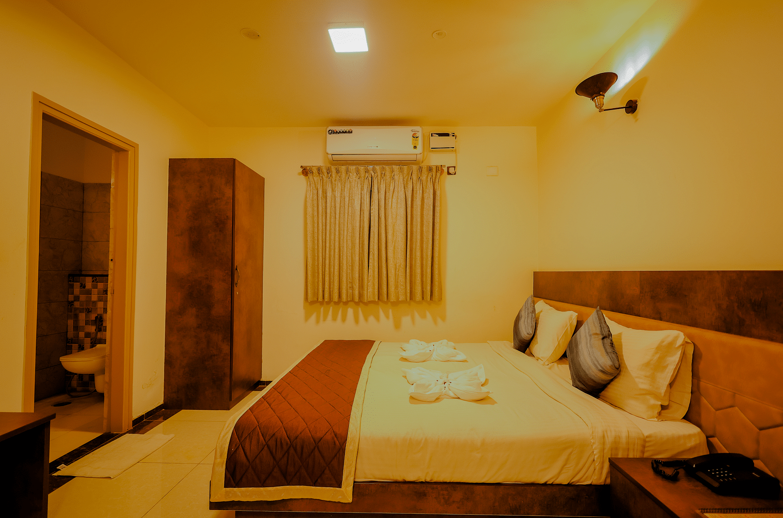 Side angle view of a room at Niratara Resort and Spa featuring a bed, window with short curtains, an almirah, and the bathroom door.