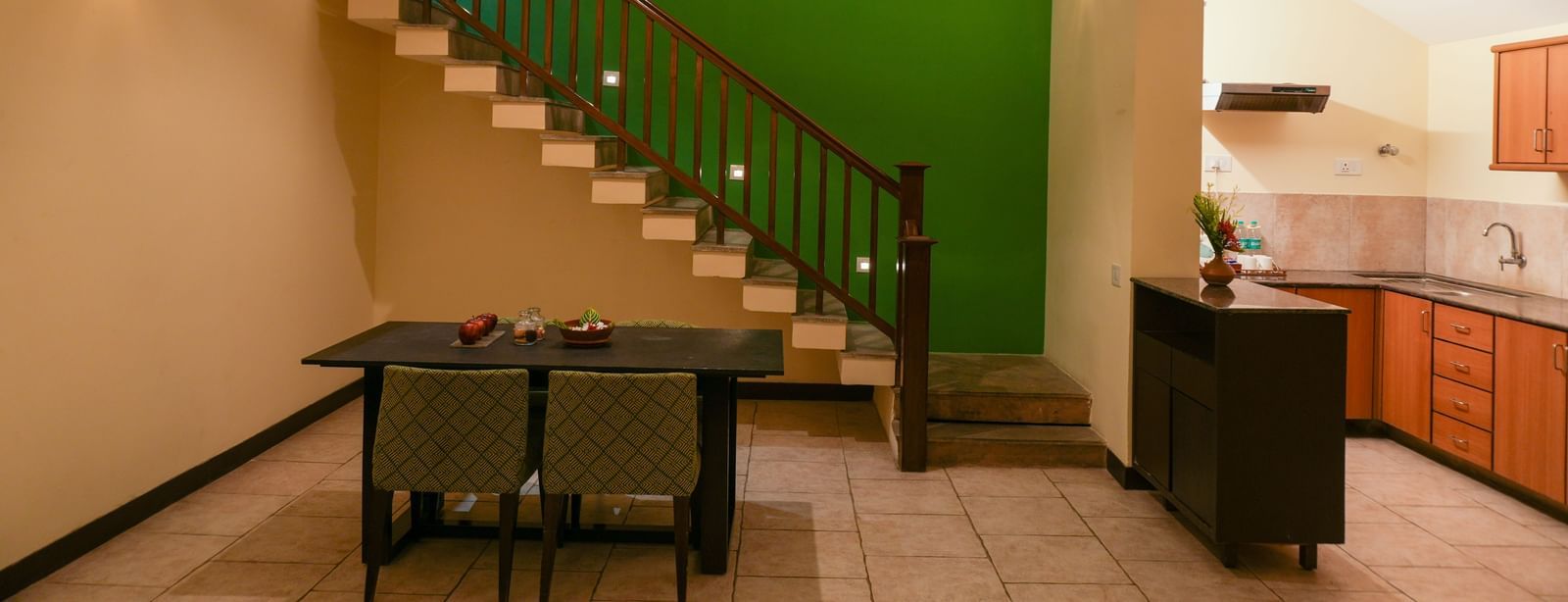 Dining area of the Retreat Villa at Vedic Village Spa Resort featuring green-painted walls, a staircase leading to the upper floor, a compact kitchen space, and an air conditioner.