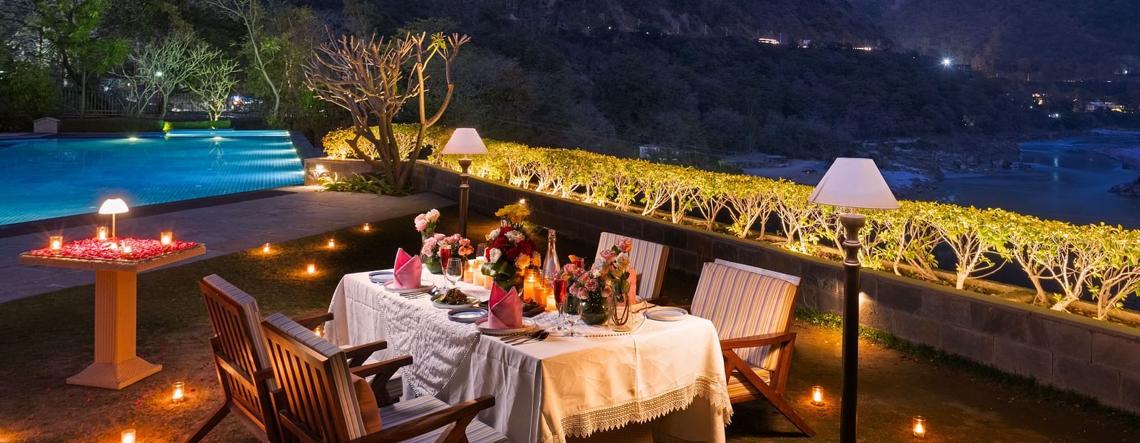 A dinner setup outdoors with a table adorned with candles and flowers,he backdrop is a beautiful mountain range at dusk.