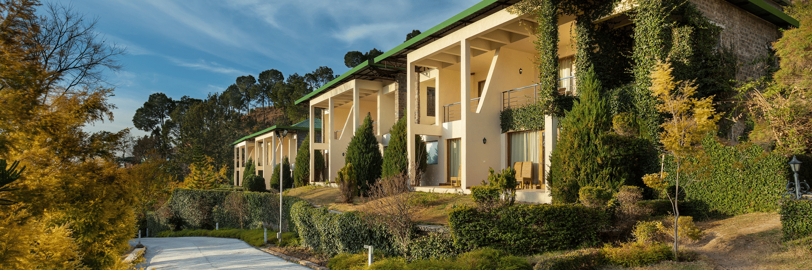 Facade of Suryavilas Luxury Resort and Spa in Solan with a paved driveway and green climbing plants on its exterior walls, under a blue sky with clouds