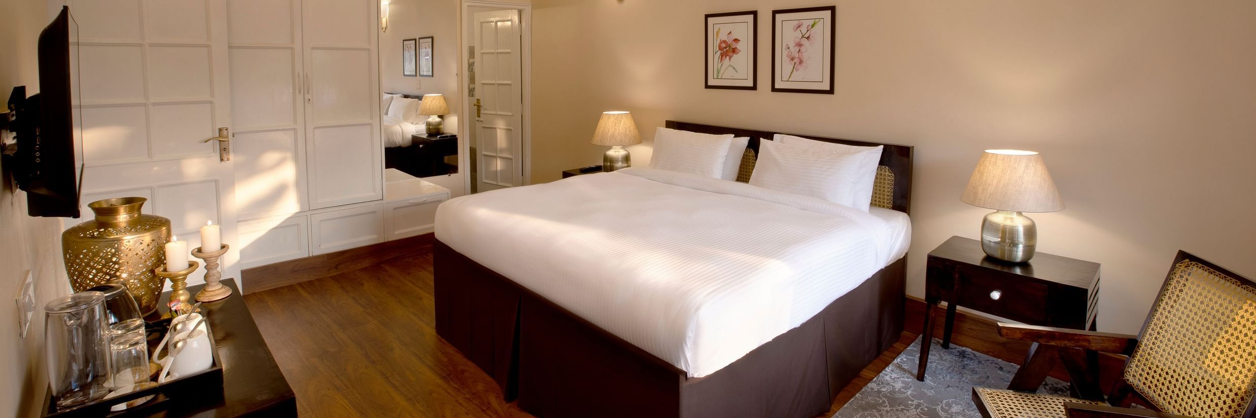 An image of super deluxe room featuring a king size bed, bedside table on either side, a flat TV screen, a table where antique items are placed, and  a chair in one corner of the room- Lamrin Boutique Cottages Rishikesh