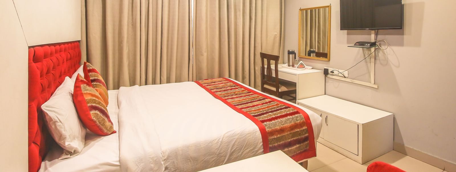 A hotel room with a plush bed, layered curtains, a TV, a desk, and a bright red accent chair, illuminated by soft ceiling lights. - Hotel Diamond Plaza, Chandigarh.