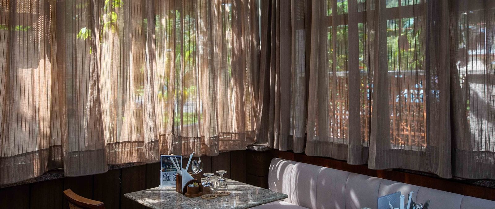 An indoor dining area at Ocean Prime Coral Reef Resort & Spa, Havelock with a wide sofa, a marble table, two glasses kept upside down on them and natural light coming through brown transparent curtains - Coral Reef Resort & Spa, Havelock