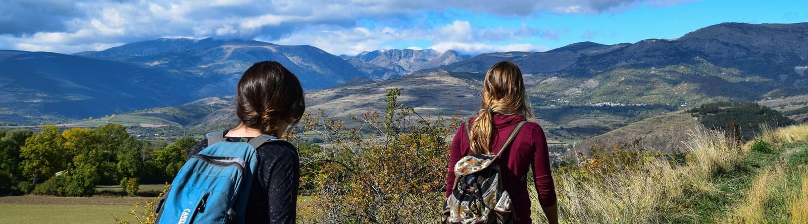 Two women with backpacks hiking along a grassy trail overlooking scenic mountains, valleys, and a partly cloudy blue sky.