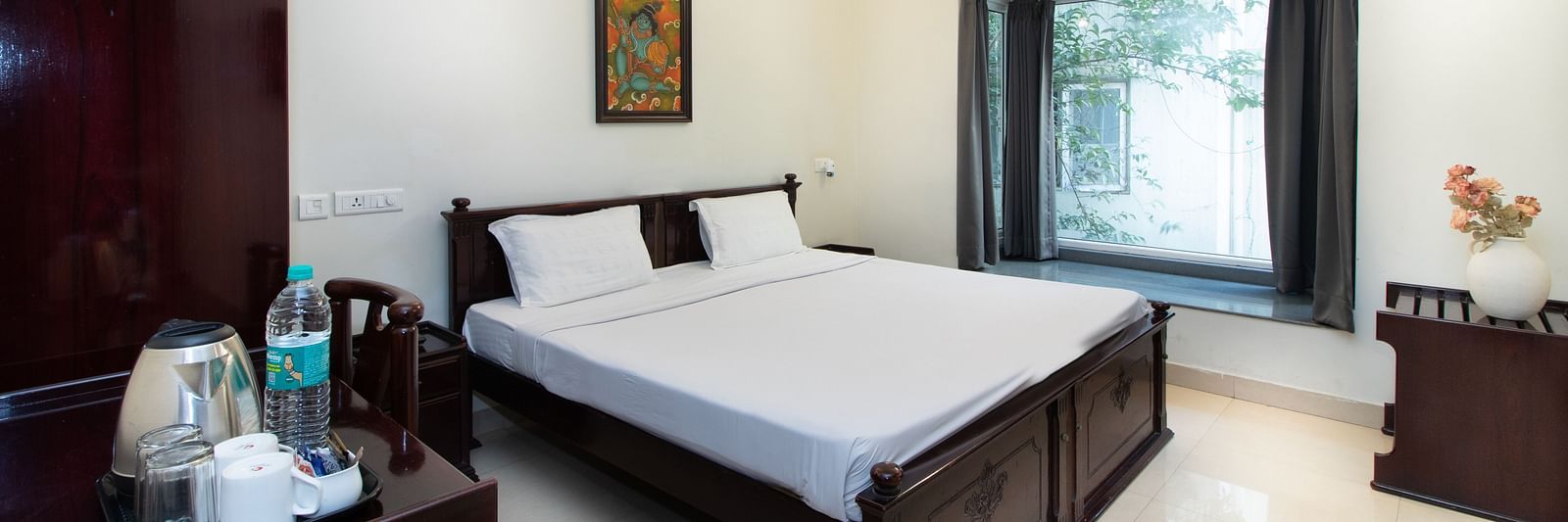 A room in Sanctum Suites Indiranagar, Bangalore, showcasing a double bed with white bedding, a bedside table with a lamp, an electric kettle placed on a table, a chair, and a window providing natural light.