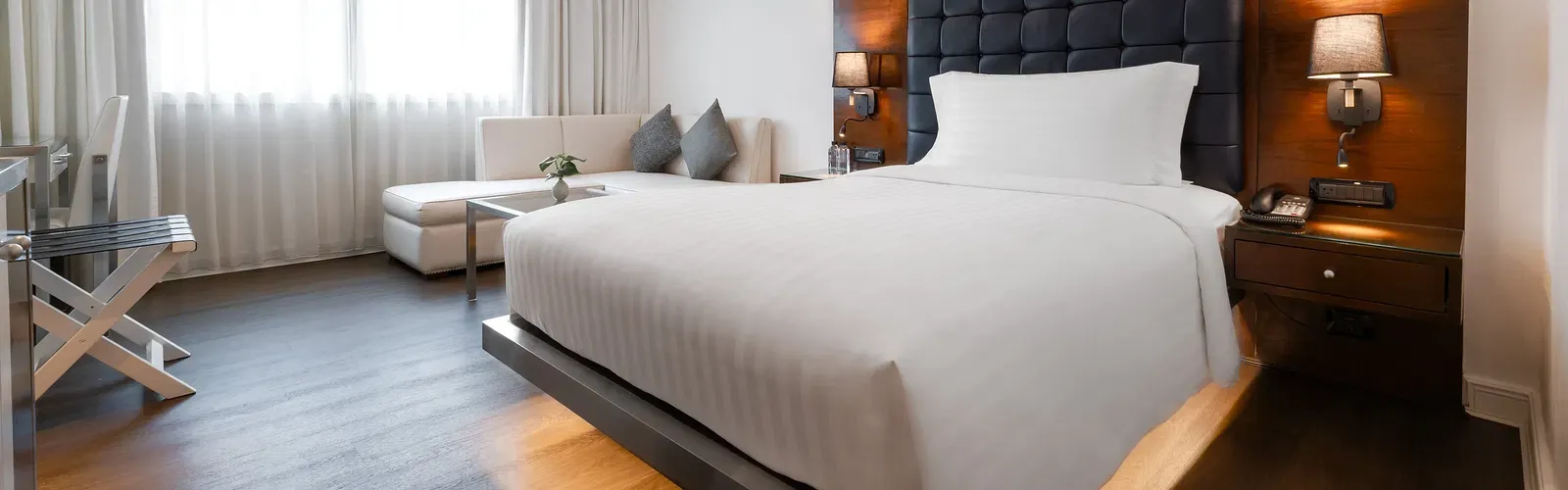 Modern hotel bedroom with tufted navy headboard, platform bed, white seating area, walnut accents, and floor-to-ceiling curtains at Night Hotel Bangkok.
