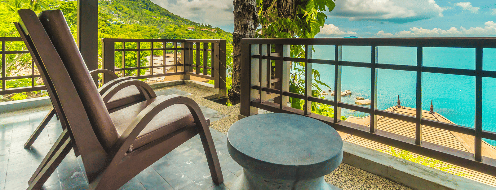 A balcony with 2 wooden lounge chairs, a small round table and a view of the sea and surrounding greenery.