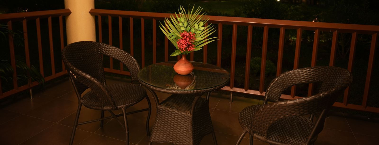 Night-time view of the balcony in the Standard Suite at Vedic Village Spa Resort featuring lounge chairs, a centre table, and wooden railings.