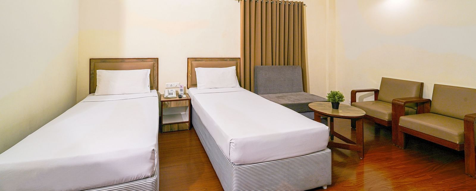 Well-lit hotel room with two single beds, wooden flooring, and a spacious seating area in Hyderabad near Ramoji Film City.