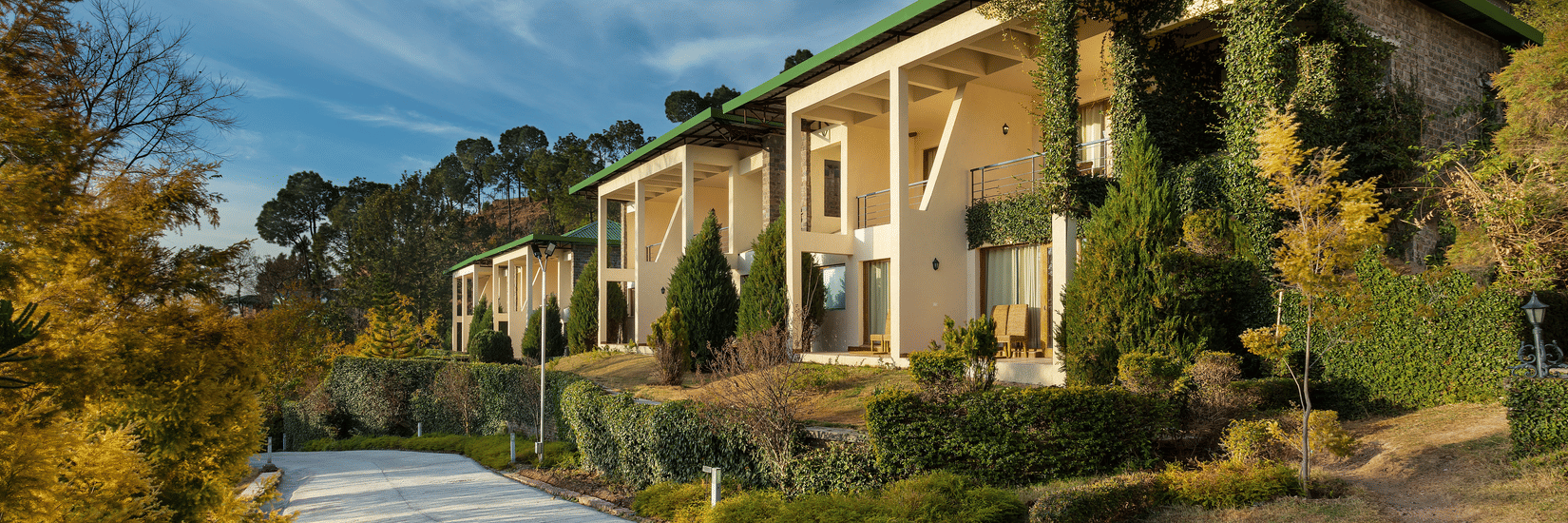Facade of Suryavilas Luxury Resort and Spa in Solan with a paved driveway and green climbing plants on its exterior walls, under a blue sky with clouds