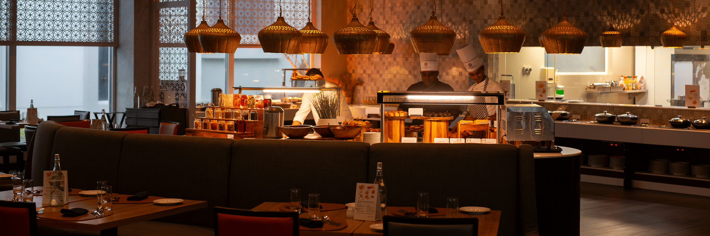  A modern restaurant interior with a buffet counter, chefs cooking, and tables set for dining. The restaurant has warm lighting and decorative hanging lamps at THE RESIDENCY TOWERS RAMESWARAM