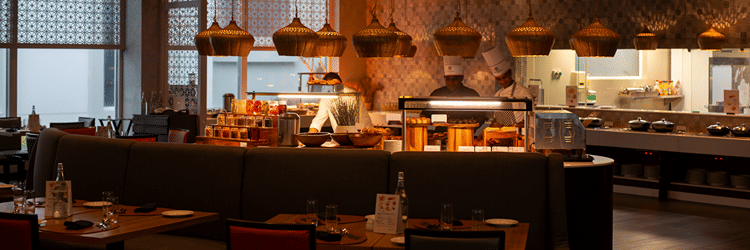  A modern restaurant interior with a buffet counter, chefs cooking, and tables set for dining. The restaurant has warm lighting and decorative hanging lamps at THE RESIDENCY TOWERS RAMESWARAM