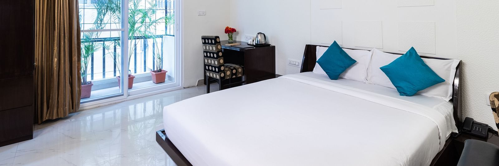A room with a double bed with blue pillows, a work desk and chair, and glass door leading to the balcony at Sanctum Suites Domlur, Bangalore.