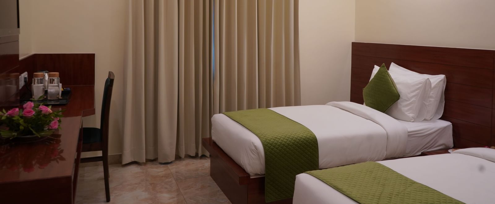 A wide shot of a hotel room with two single beds. The beds have white comforters with green accents and are separated by a small table.
