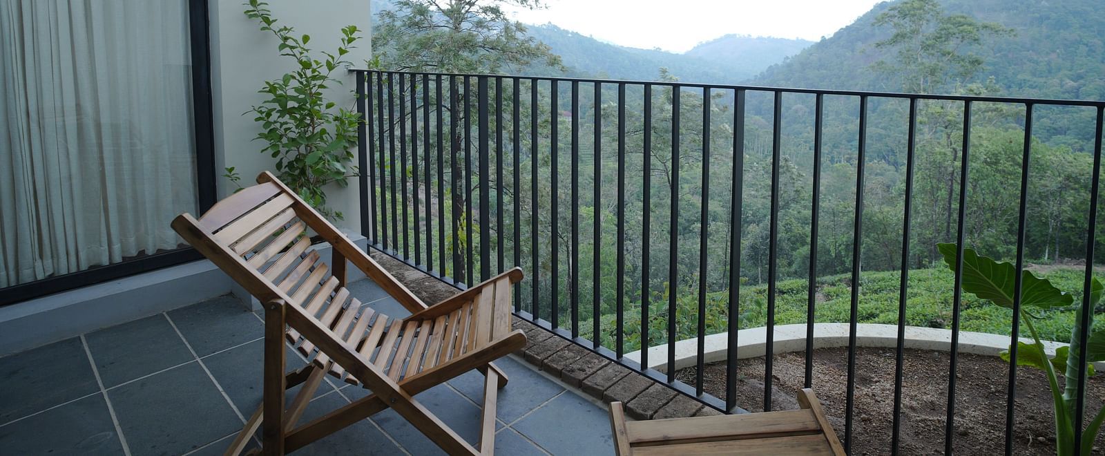  A serene balcony at Vaasta Vagamon with a wooden lounge chair, offering expansive views of lush green mountains and valleys.
