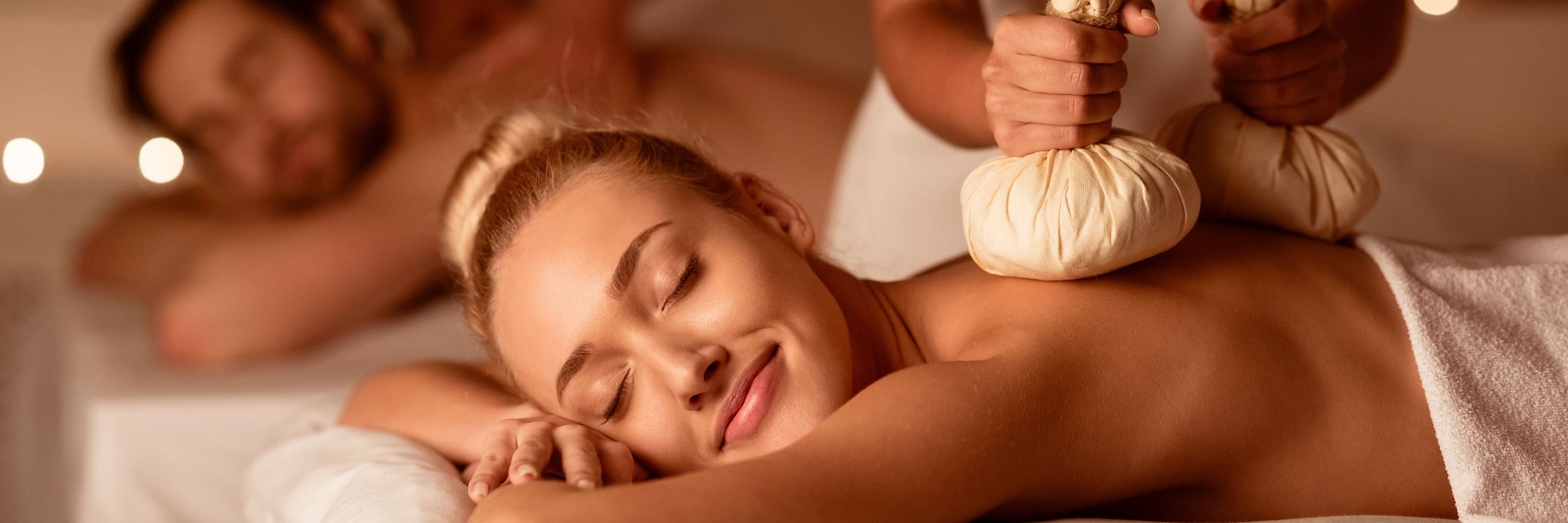 A couple undergoing Ayurvedic kizhi massage, laying on white beds, assisted by two practitioners. 