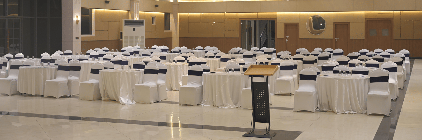 A front-angled view of the banquet hall set with round tables and white seating under warm ceiling lights at Hotel Sonar Bangla Mayapur.