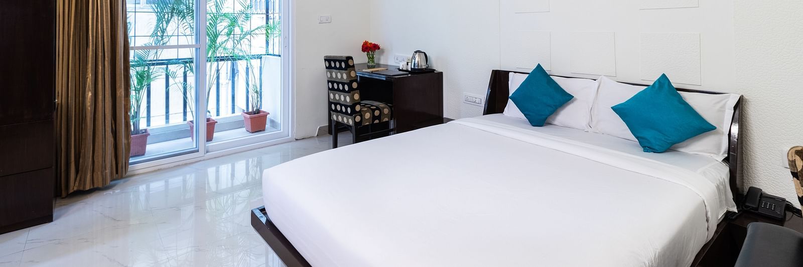 A room with a double bed with blue pillows, a work desk and chair, and glass door leading to the balcony at Sanctum Suites Domlur, Bangalore.
